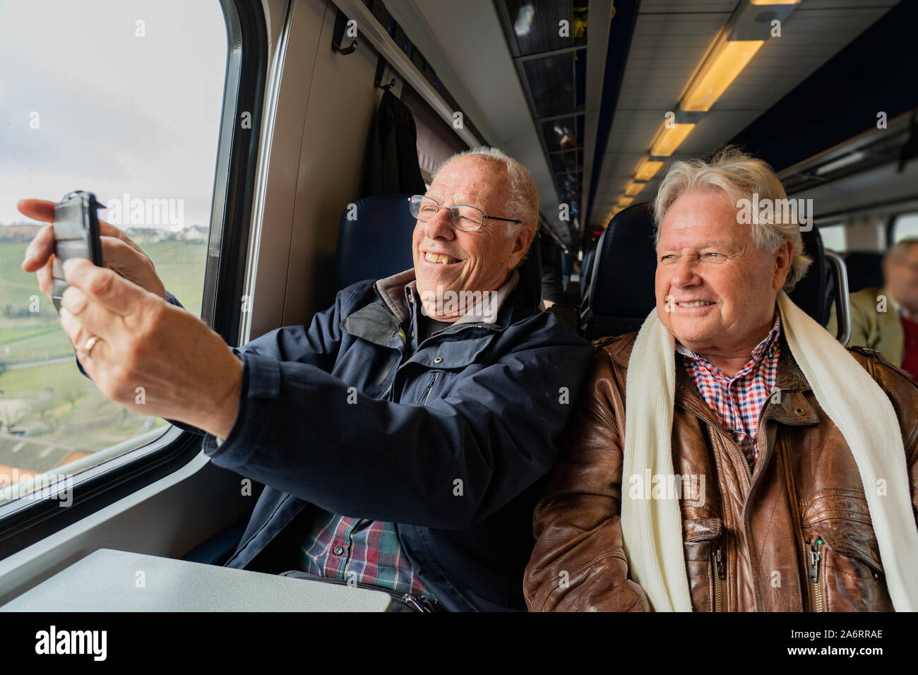 Older man with a good friend in casual wear is taking a self portrait with a smart phone in train in Switzerland at daylight. Stock Photo