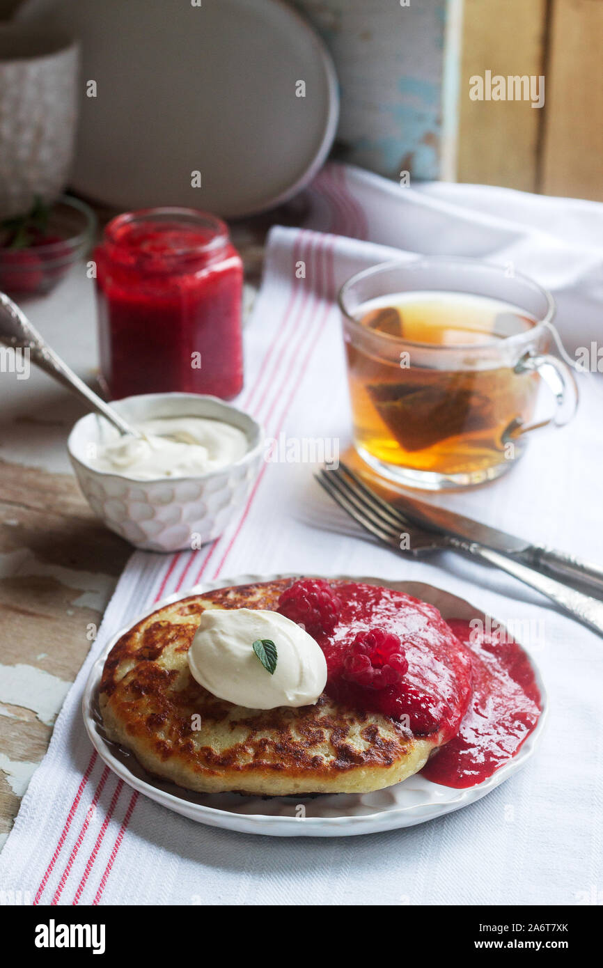 Curd pancakes with strawberry sauce, sour cream and raspberries, and herbal tea on a light table. Rustic style. Stock Photo