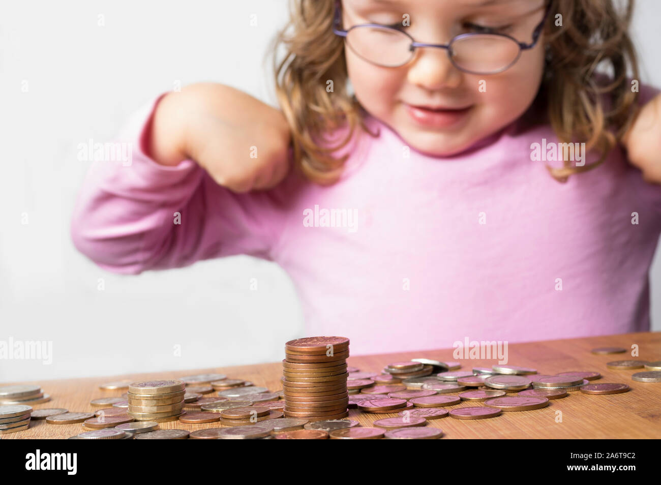 Smiling girl wearing glasses dressed in pink counting coins for savings. Focus on the coins at the front. Clean neutral background and copy space on t Stock Photo