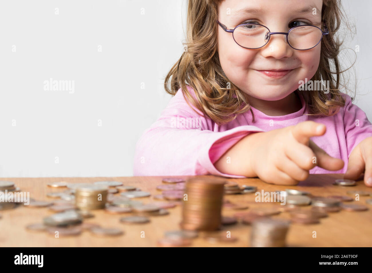 Smiling girl wearing glasses dressed in pink counting coins for savings. Focus on the face. Clean neutral background and copy space on the left. Stock Photo