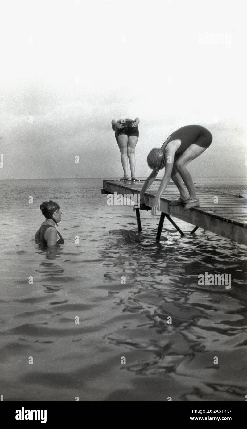 1920s, historical, outside in a large of water, possibly a lake, three female swimmers in the bathing wear of the era having fun diving off a wooden jetty on wheels, England, UK. Stock Photo