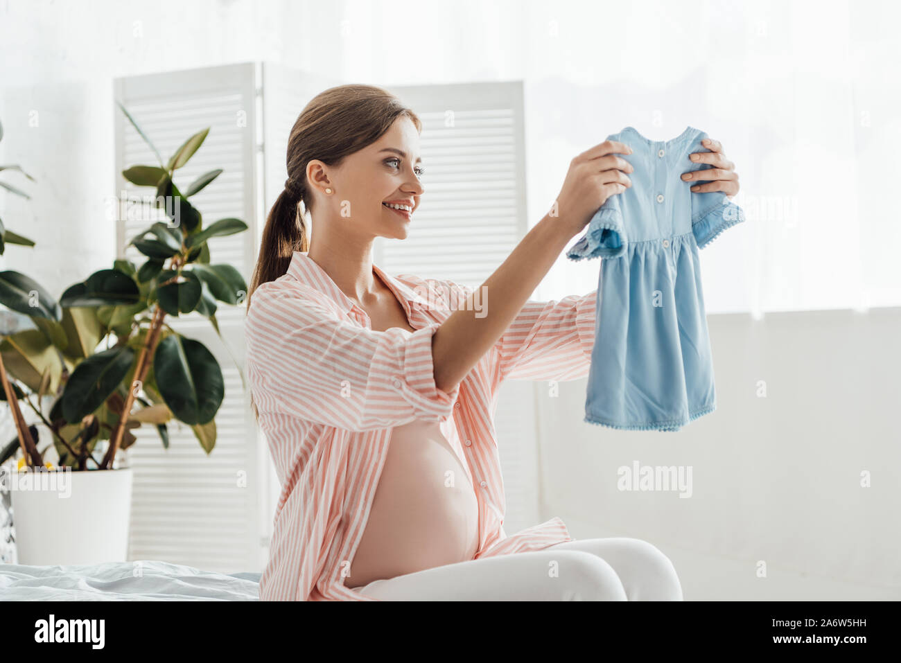 happy smiling pregnant woman sitting on bed and holding baby clothes Stock Photo