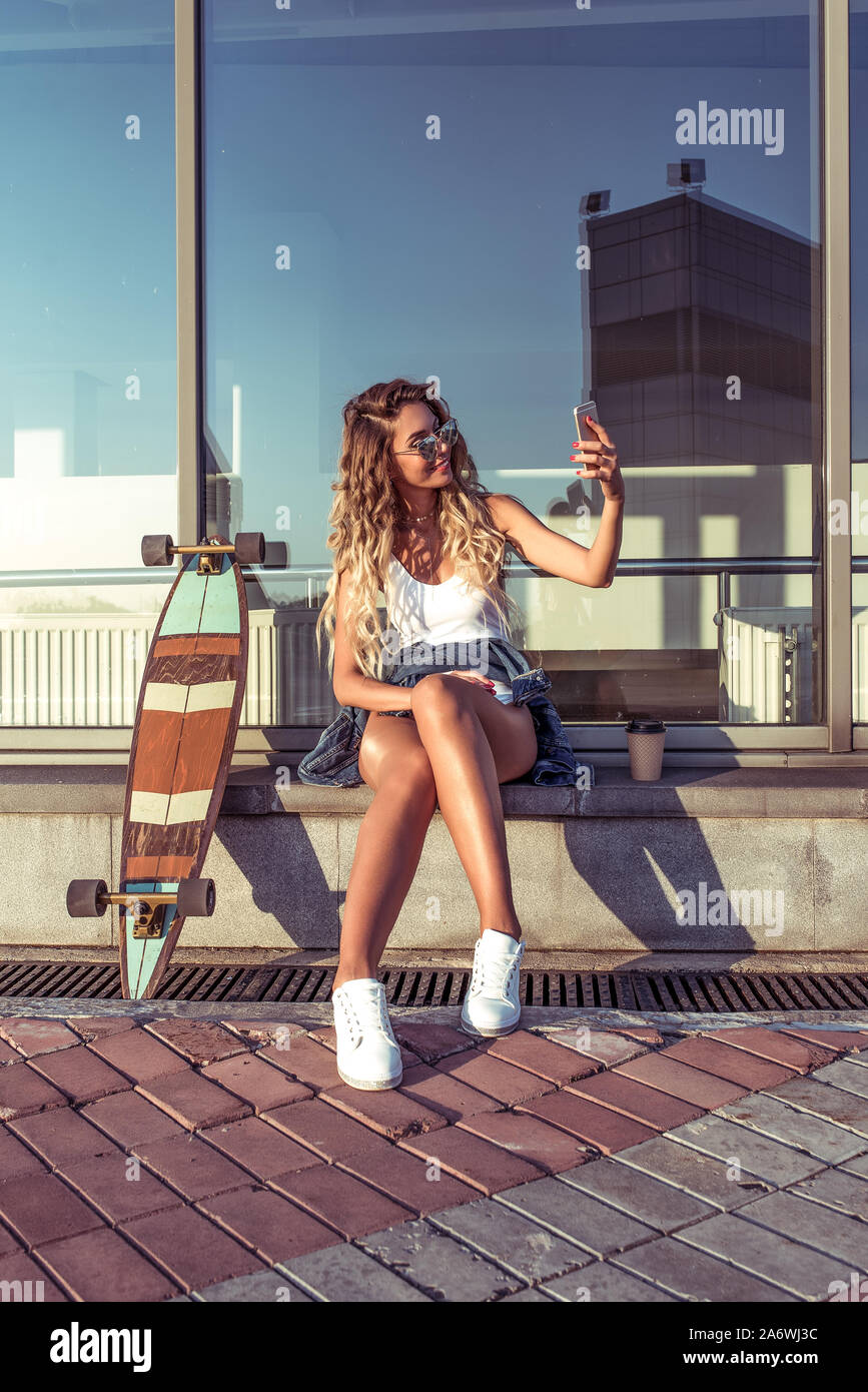 beautiful girl in city, sits makes video call, cellphone selfie on phone,  skateboard board longboard. Tanned figure, long hair sunglasses, glass  Stock Photo - Alamy