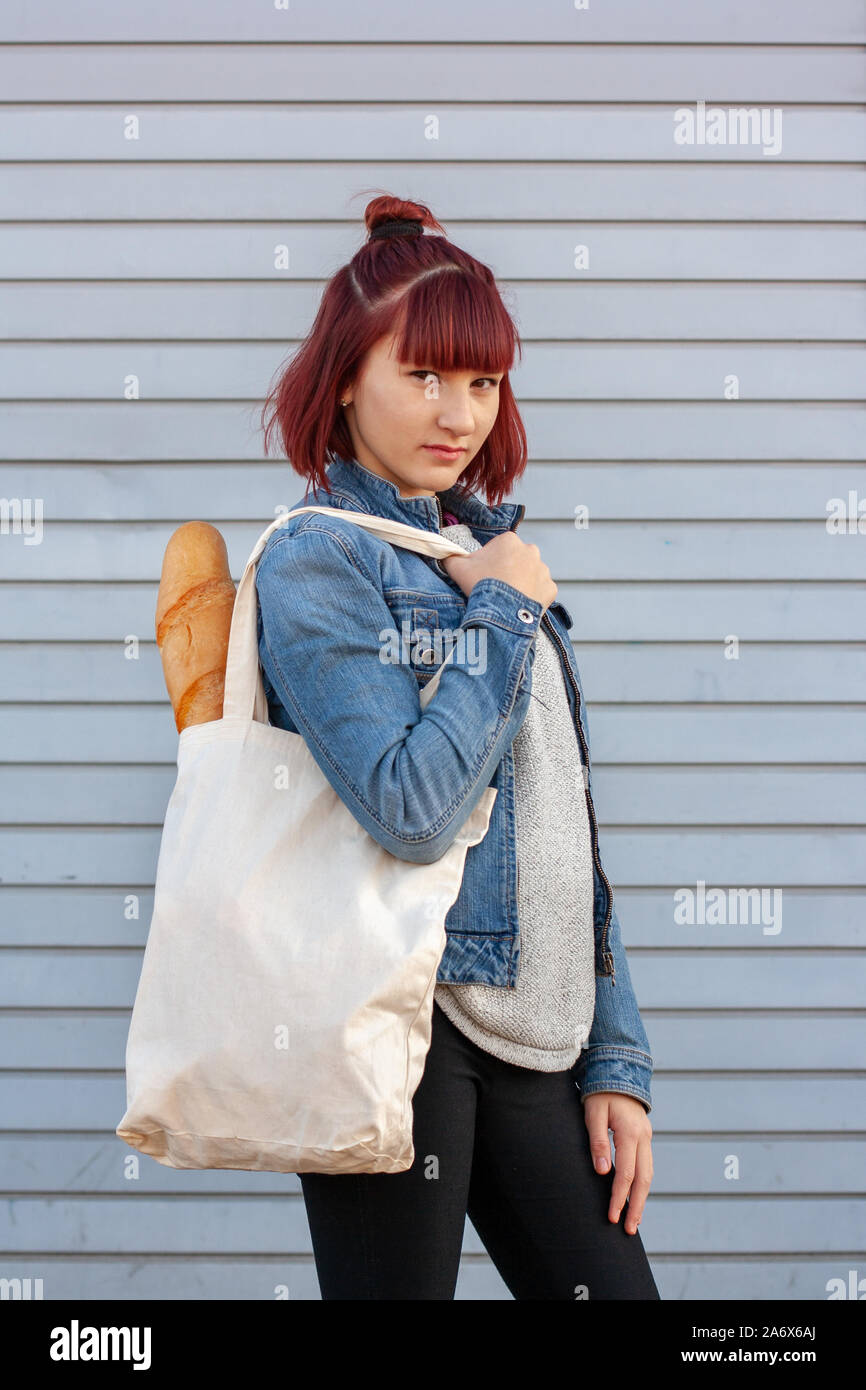 Long french bread in an eco textile bag on the shoulder of a young girl with red hair. Girl in a denim jacket and black pants. Stock Photo