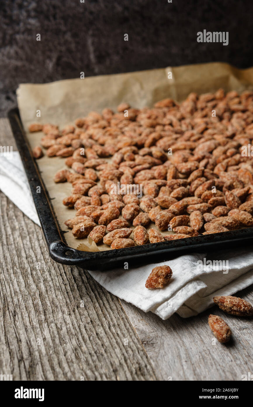 Burnt almonds outspread on black baking tray, paper, white cloth on rustic textured wooden table, three burnt almonds laying in foreground Stock Photo