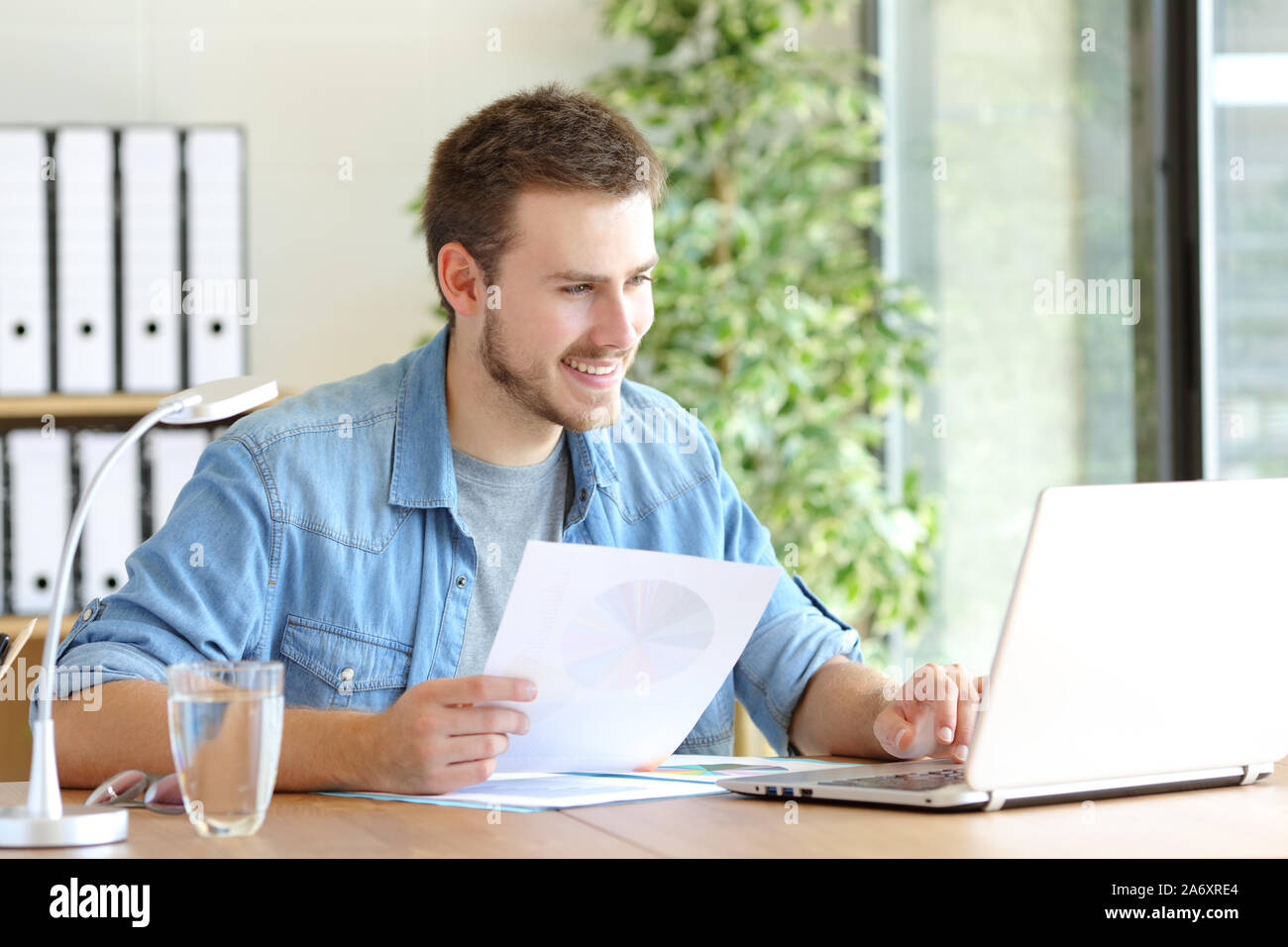 Casual happy entrepreneur working using laptop holding document comparing data at office Stock Photo