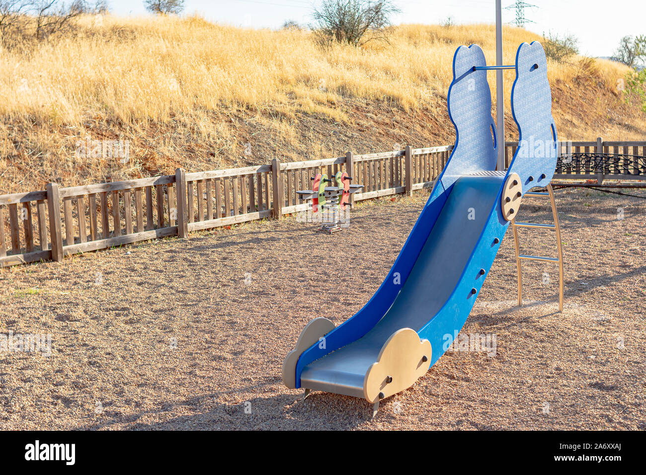 Metallic and wooden slide with other swings in a children's playground with gravel and wooden fencing for child safety Stock Photo