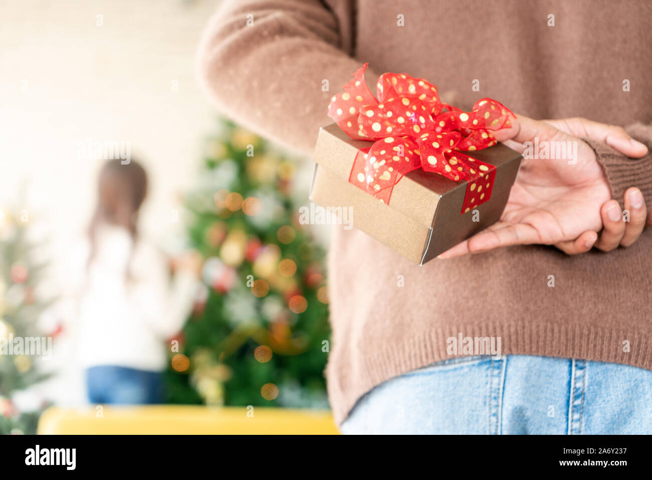 Man hold christmas gift behind his back for surprise his girlfriend while she decorating christmas tree in Chgristmas holiday season greeting. Stock Photo