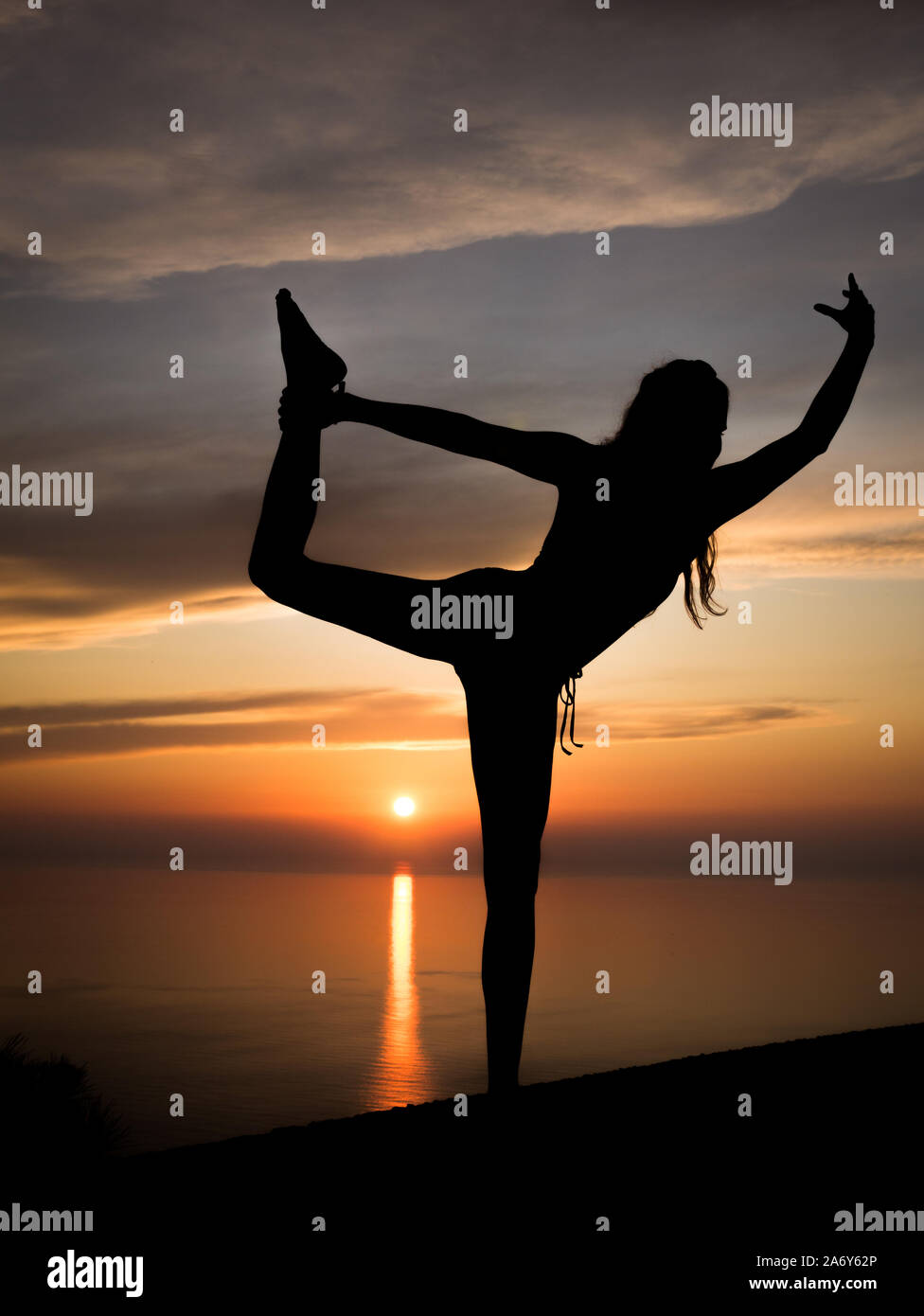 Silhouette of young and sportive woman practicing yoga (standing scale pose) with romantic sunset and ocean in the background at golden hour Stock Photo