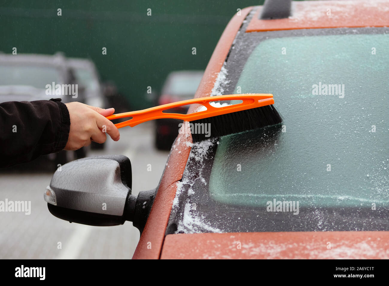 Brush in mans hand. Man clears snow from icy windshield of orange car. Man is brushing automobile window. Stock Photo
