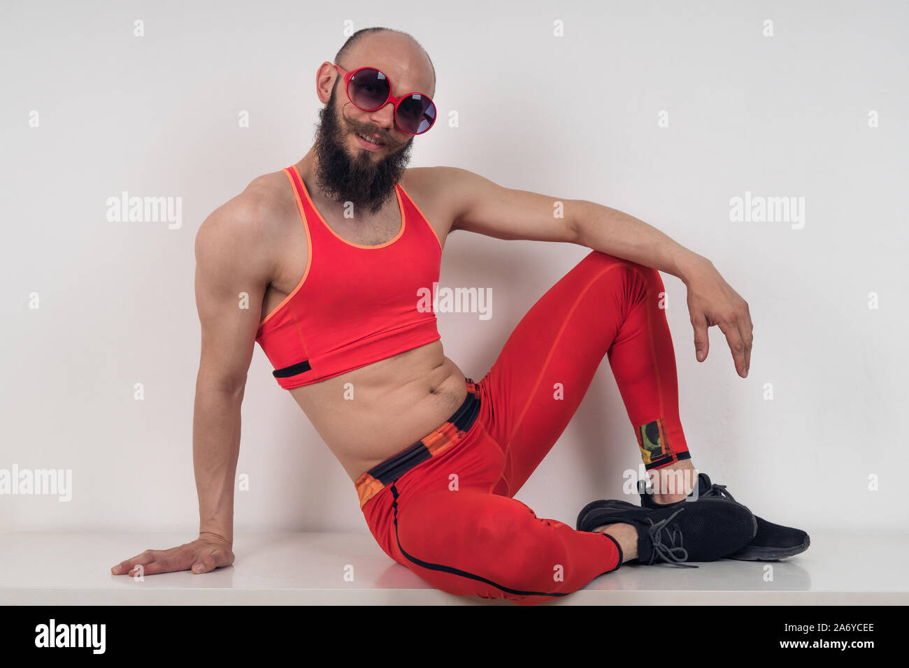 Funny guy in tight red tracksuit and sunglasses posing sitting. Stock Photo