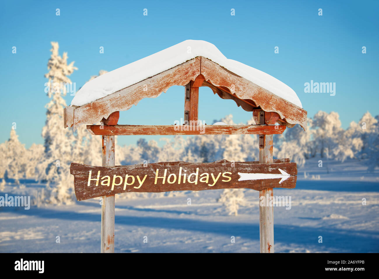 Happy holidays written on a wooden direction sign, blue sky and winter snowy tree landscape background seasons greetings christmas card Stock Photo