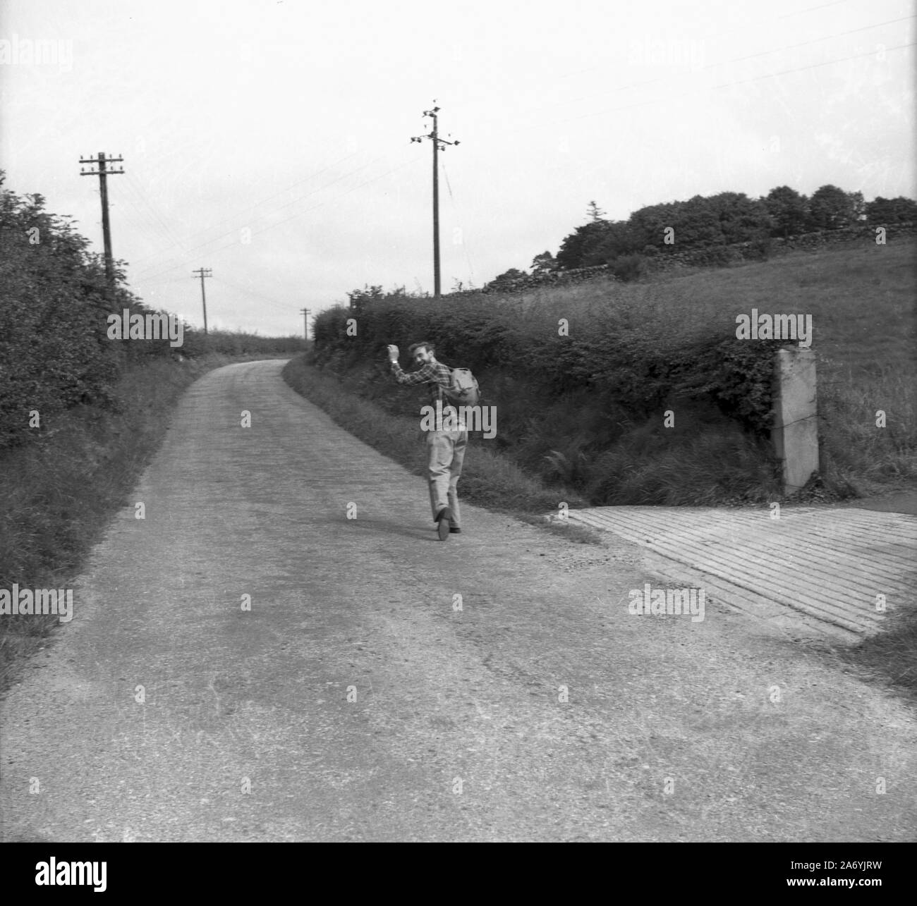 1950s, historical, a male walker, with rucksack, on a hedge lined rural road, looking back over his shoulder, hand in the air, waving good bye, as he head  up the road, England, UK. Stock Photo