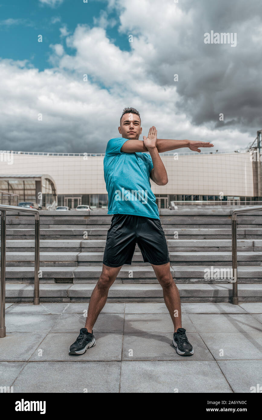 Athletic man, summer afternoon training in city, active lifestyle, modern fitness workout, sportswear t-shirt shorts sneakers. Warming up the muscles Stock Photo