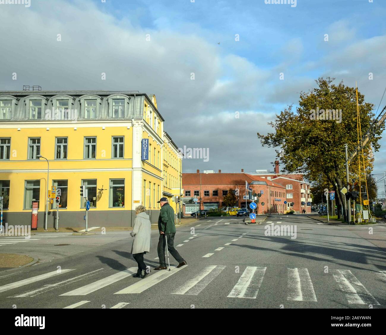 An elderly couple on a pedestrian crossing in front of a bright yellow hotel in Ystad, Sweden. Stock Photo