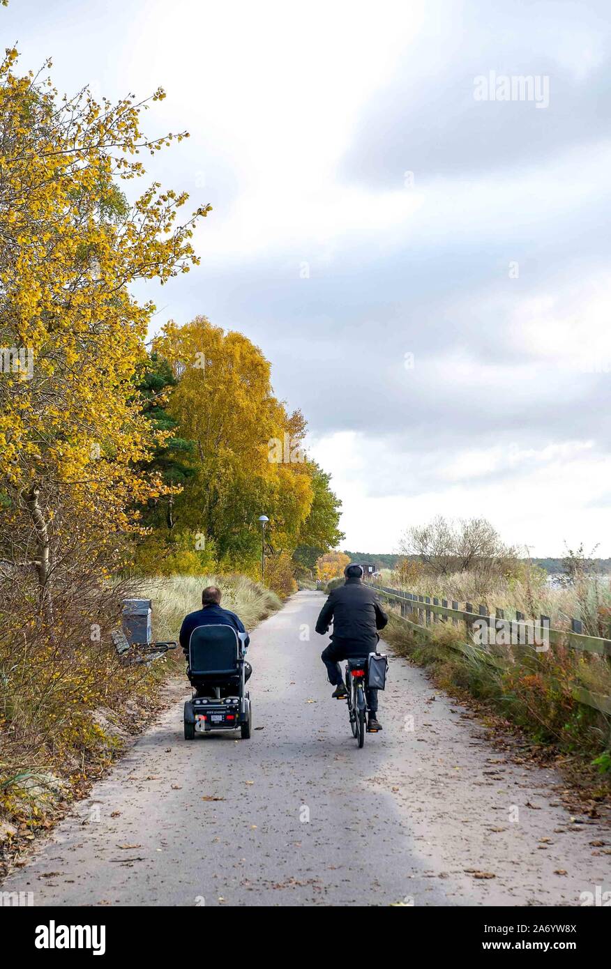 Two friends ride side-by-side, one on a bicycle and one on a mobility scooter, on the paved path along the seaside in Ystad, Sweden. Stock Photo