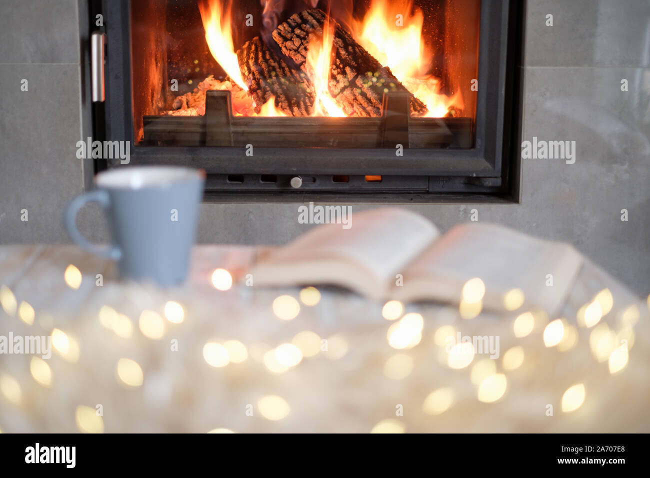 Open book, cup of tea and luminous garland near burning fireplace. Hygge concept Stock Photo