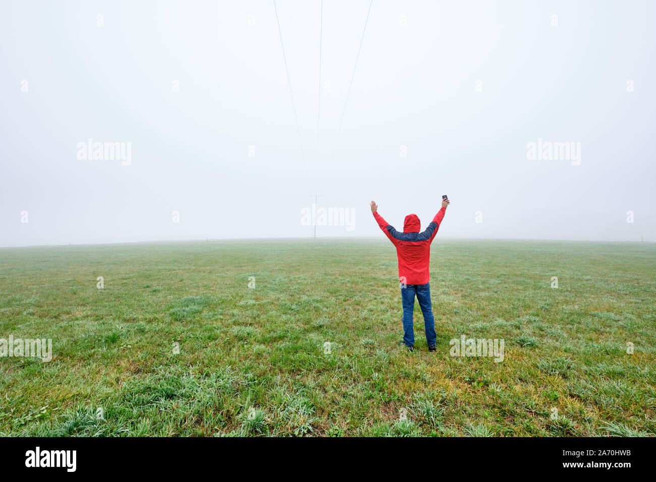 Rear view of man in red jacket standing on a green meadow and raising his arms in joy in front of a foggy nowhere landscape. Seen in October in German Stock Photo