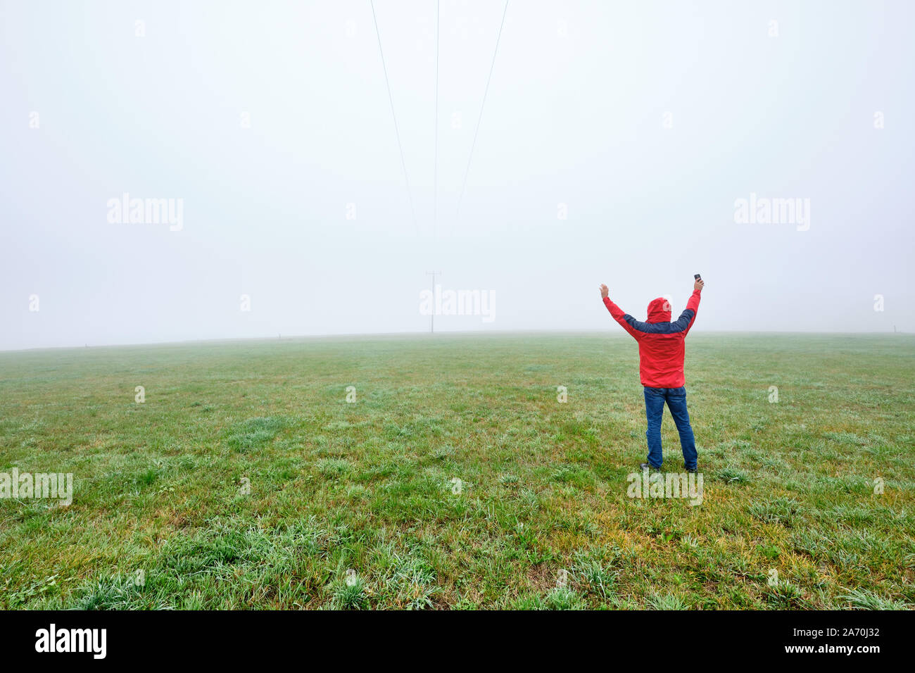 Rear view of man in red jacket standing on a green meadow and raising his arms in joy in front of a foggy nowhere landscape. Seen in October in German Stock Photo