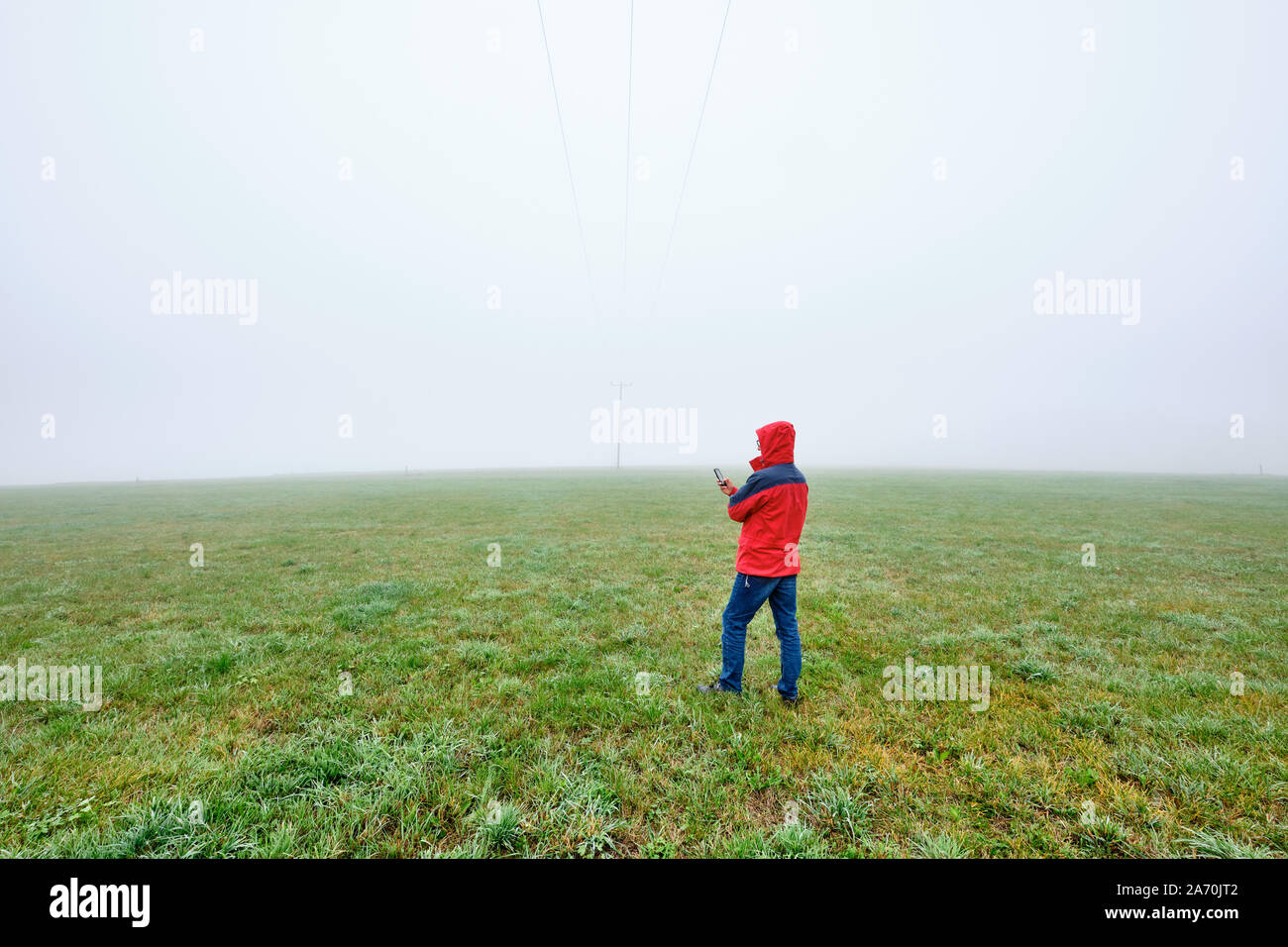 Rear view of man in red jacket standing on a green meadow and looking on his smartphone in front of a foggy nowhere landscape. Seen in October in Germ Stock Photo