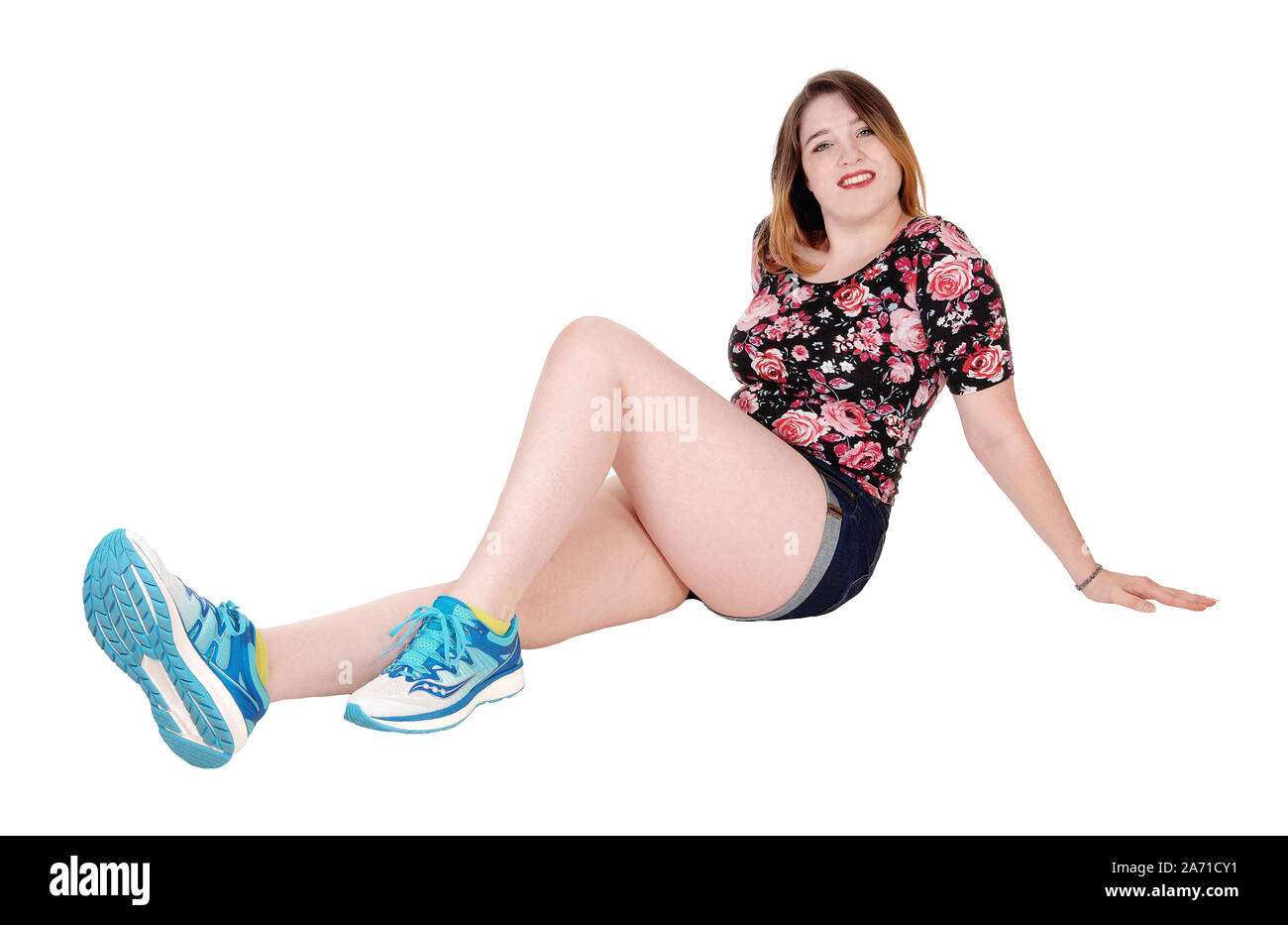 A young beautiful woman sitting in shorts and sneakers on the floor, looking into the camera, isolated for white background Stock Photo