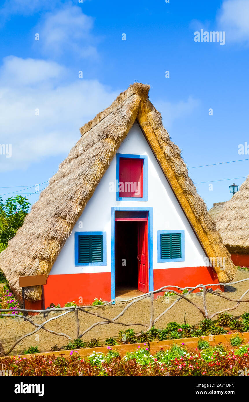 Traditional house in Santana, Madeira island, Portugal. Small, wooden, triangular houses with thatched roof and colorful facade. Part of Portuguese heritage. Flower garden in foreground. Sunny day. Stock Photo