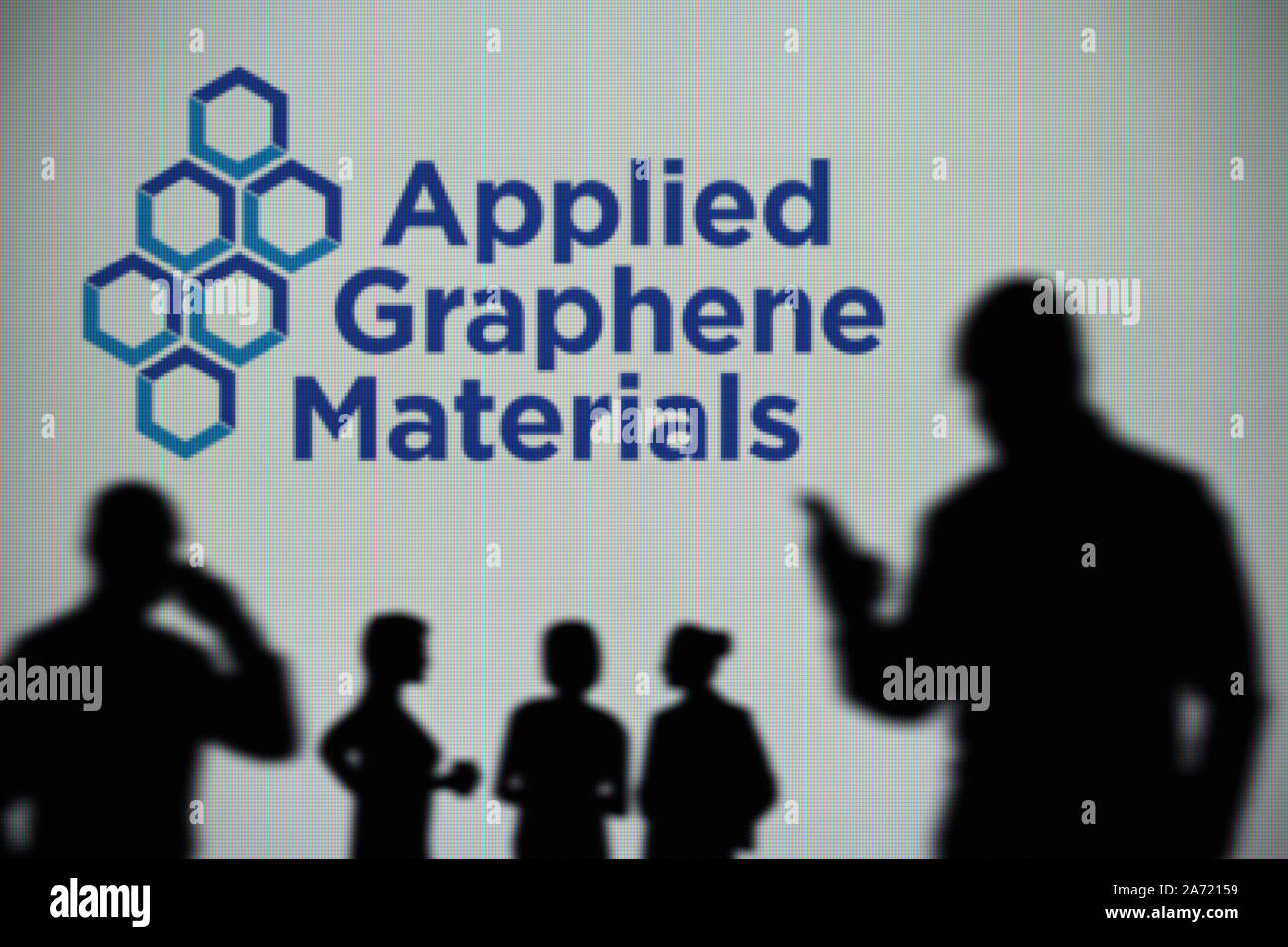 The Applied Graphene Materials logo is seen on an LED screen in the background while a silhouetted person uses a smartphone (Editorial use only) Stock Photo