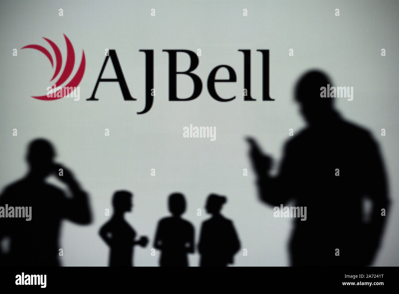 The AJ Bell logo is seen on an LED screen in the background while a silhouetted person uses a smartphone (Editorial use only) Stock Photo