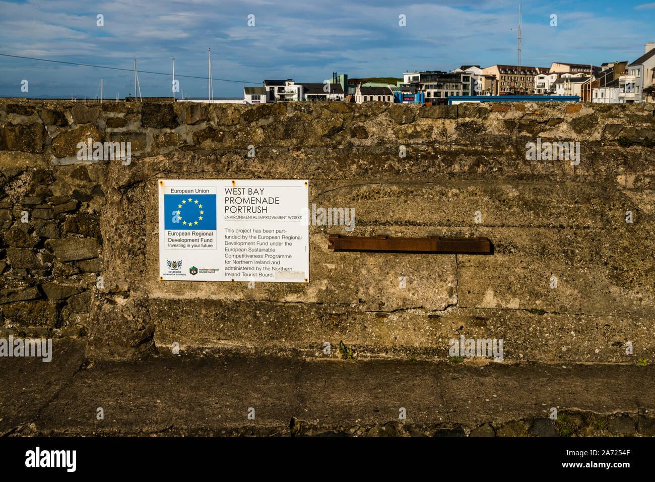 A sign from the European Regional Development Fund marks environmental improvement works on the historic stone wall of the West Bay Promenade in Portrush, County Antrim, Northern Ireland. The funding ran under the "European Sustainable Competitiveness Programme for Northern Ireland" and was administered by the Northern Ireland Tourist Board. The skyline of the coastal town of Portrush stretches out in the background. Stock Photo