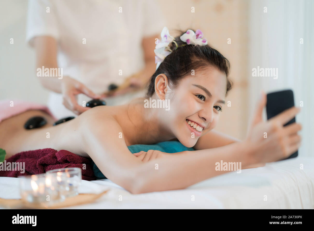 Beautiful young Asian woman smile and holding smartphone receiving rock therapy massage taking selfie in spa salon. Hand putting rock on female back, Stock Photo