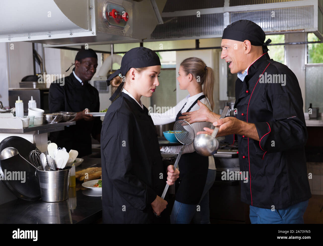 Enraged chef expressing dissatisfaction with work of frustrated girl in restaurant kitchen Stock Photo