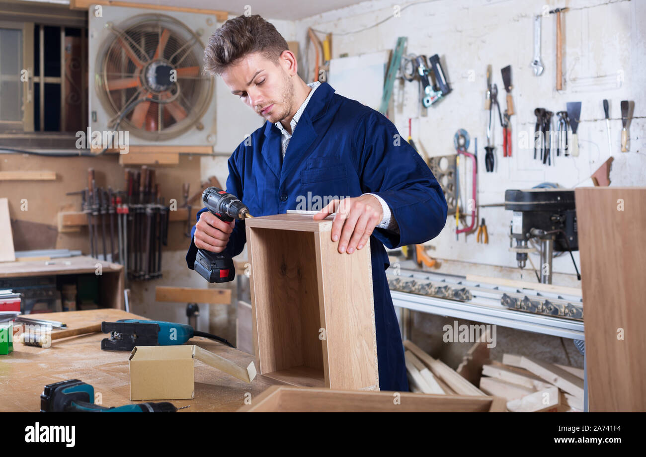 Young positive woodworker using drilling machine to make drawer at workshop Stock Photo