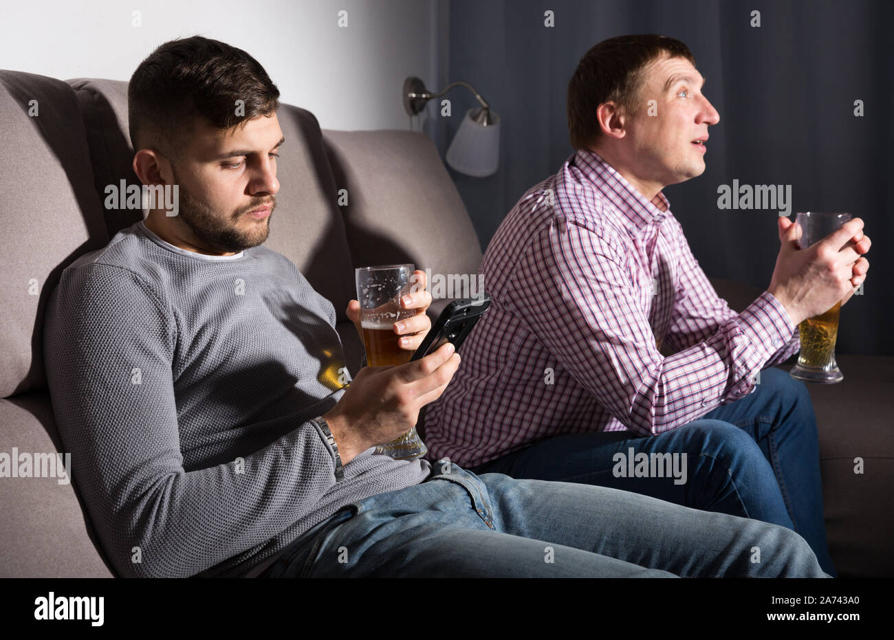 Bored guy sitting on sofa at home with phone while friend watching tv ...