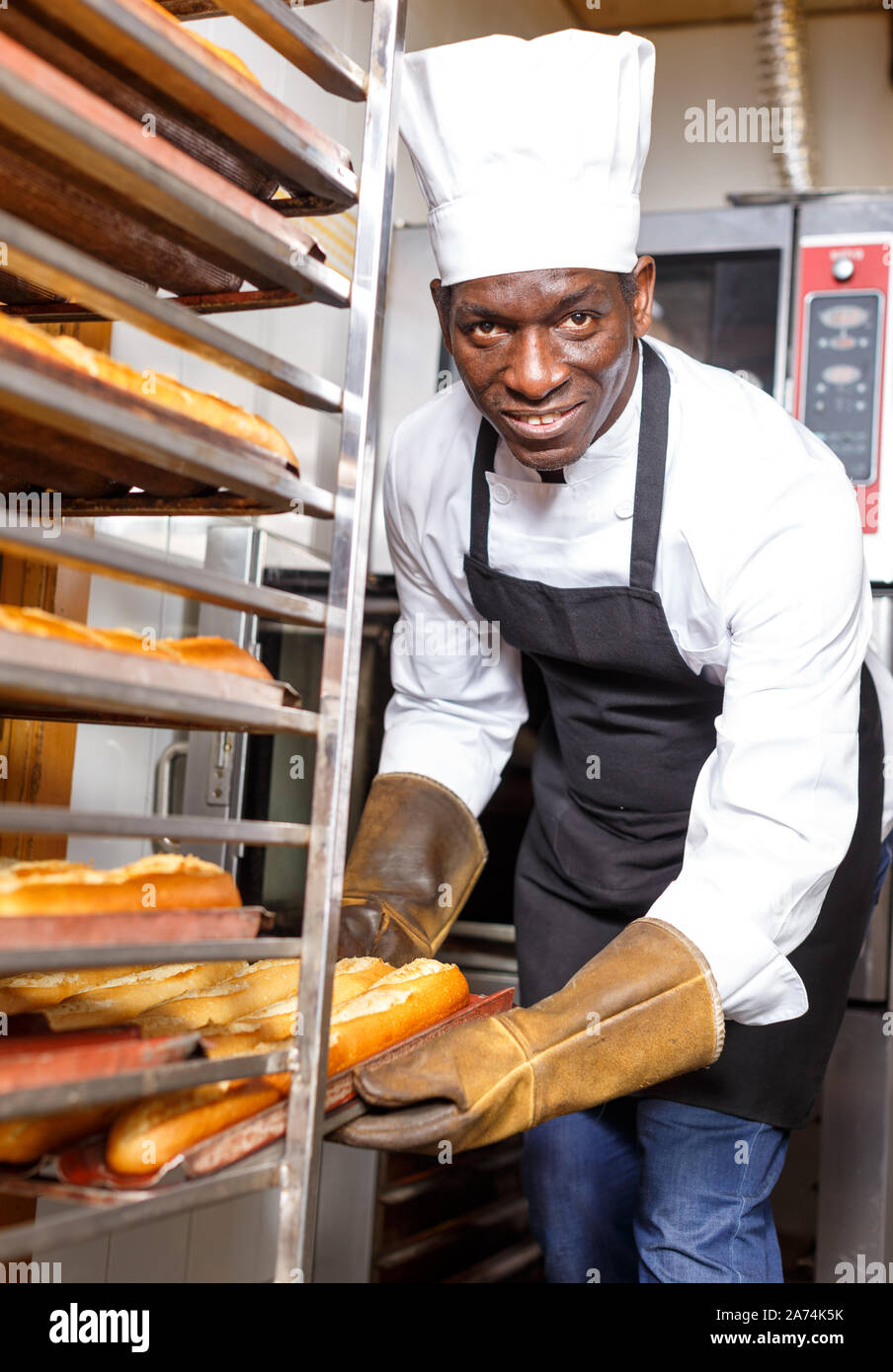 Confident worker of bakery wearing uniform and heat resistant gloves putting hot baked baguettes on tray rack Stock Photo