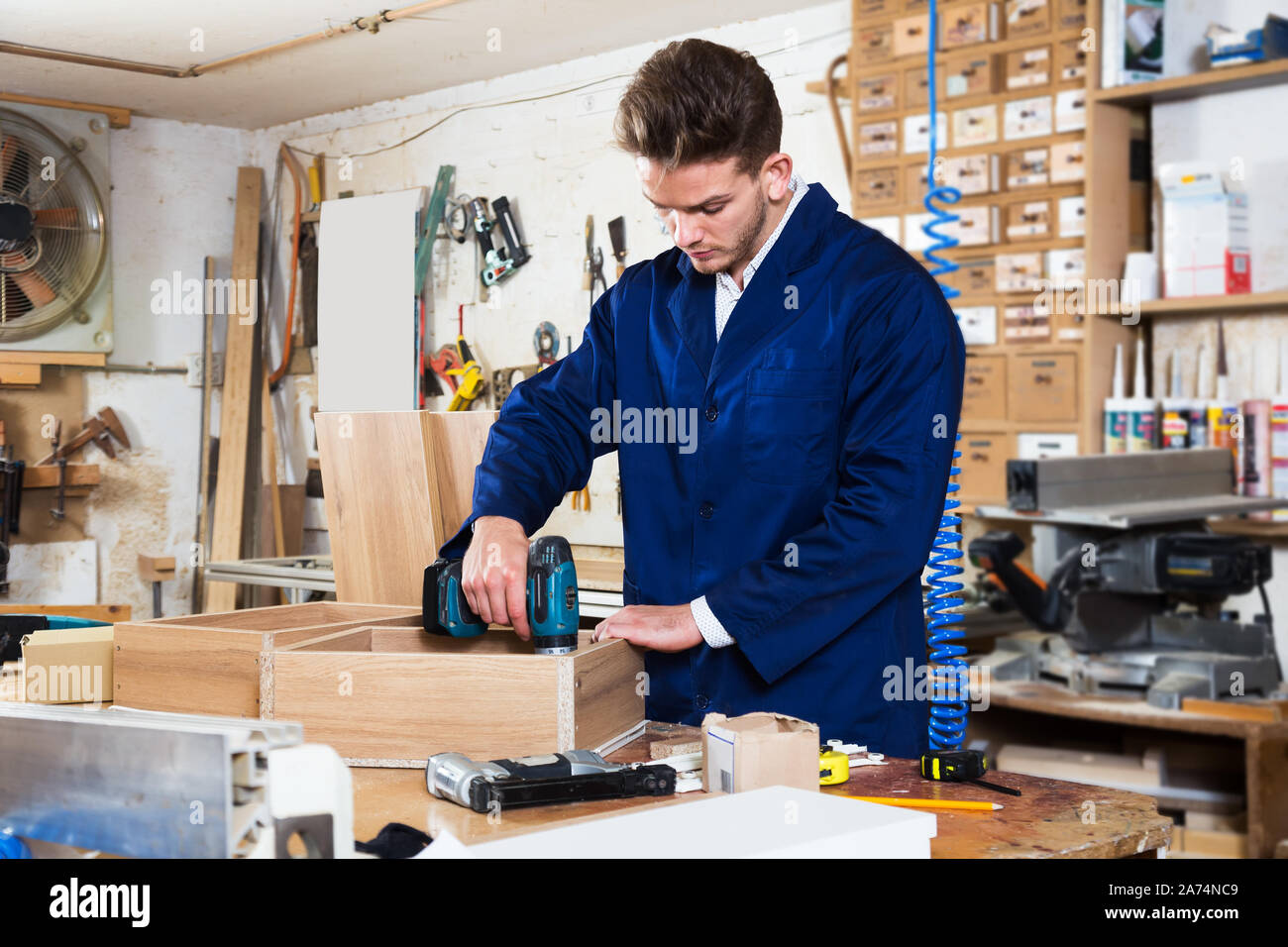 Young positive man using drilling machine to make drawer at workshop Stock Photo