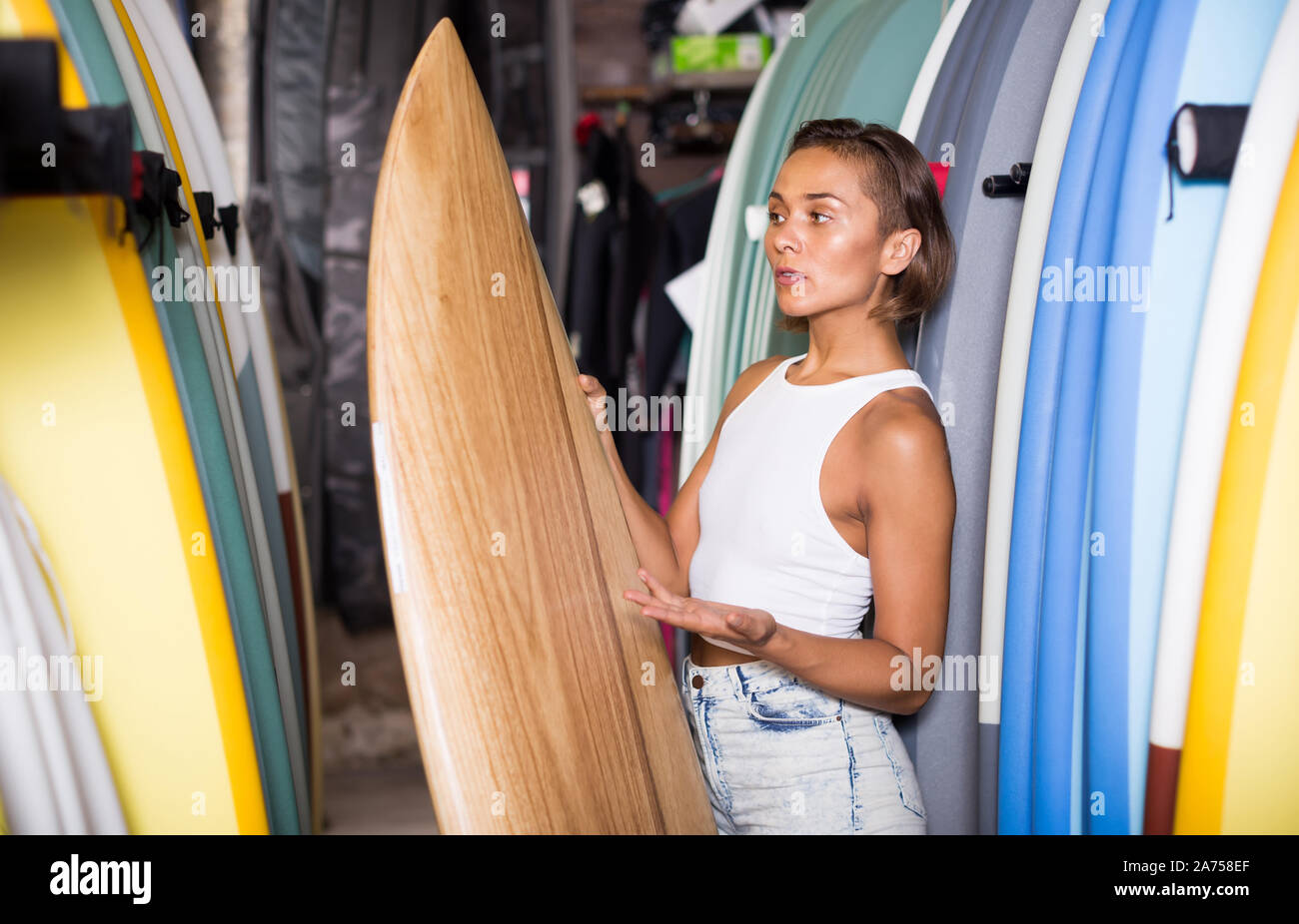 Positive spanish female holding surfboard in the sports store Stock