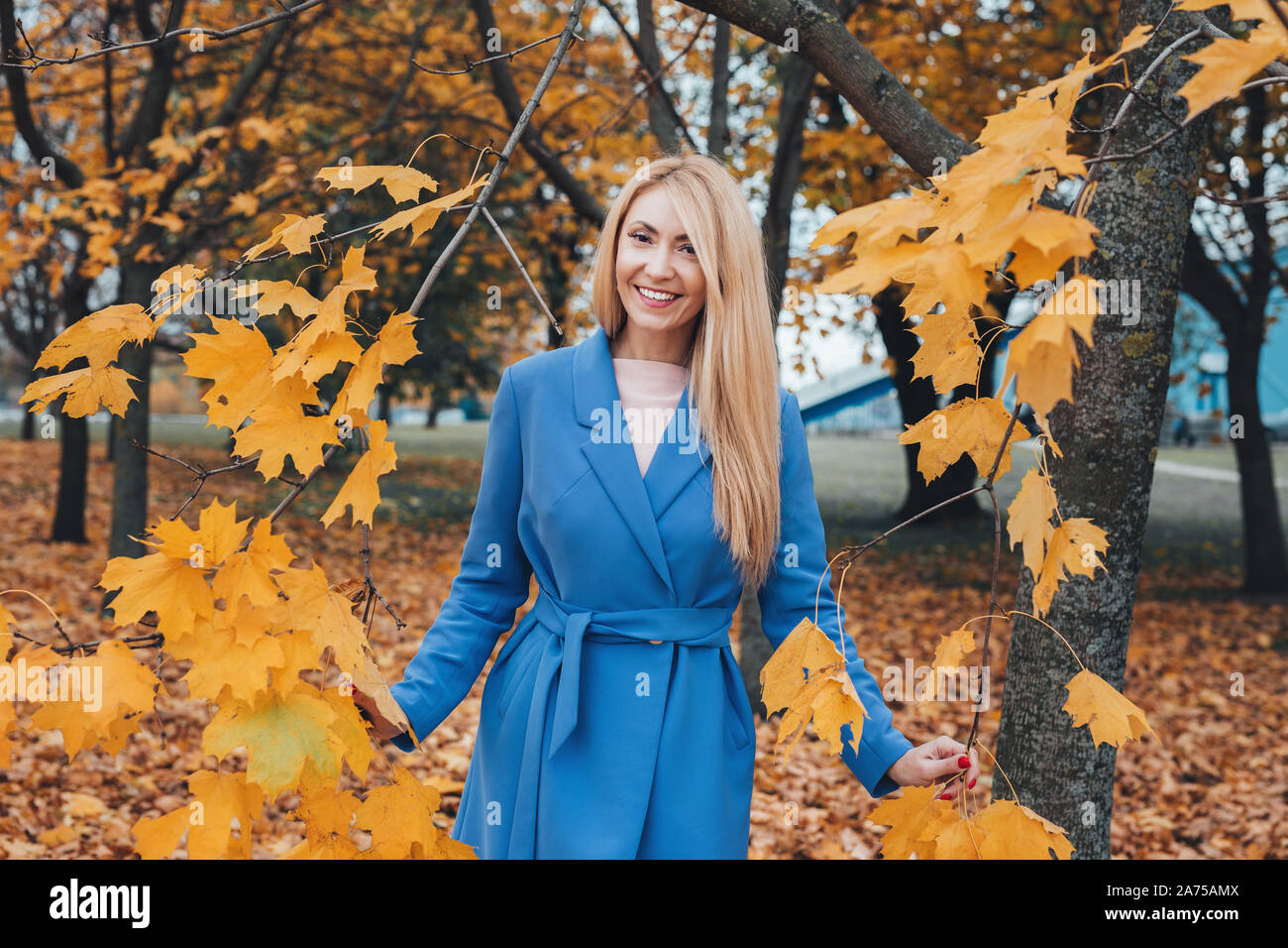 Smiling blonde mature woman in blue coat looking at camera in autumn park Stock Photo