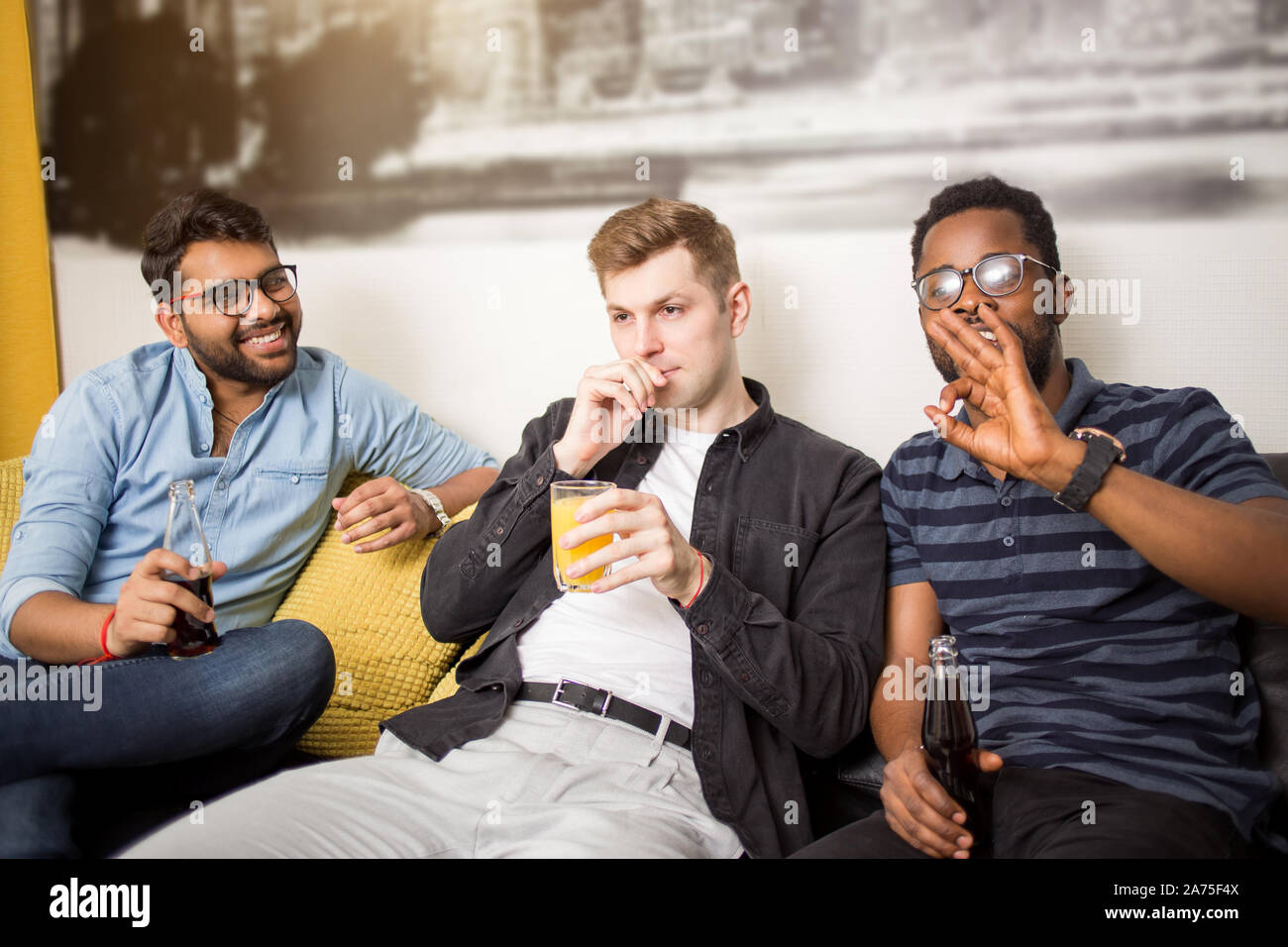 Portrait of three smiling male friends sitting on couch with nonalcoholic drinks looking to camera and showing ok gesture. Football fans watching socc Stock Photo