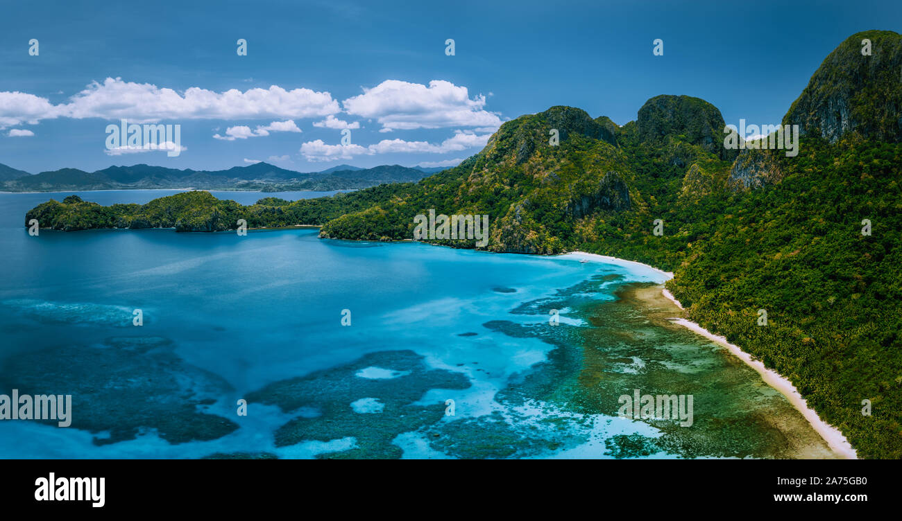 Aerial drone panoramic view of uninhabited tropical island with rugged mountains, rainforest jungle and big blue bay with shallow ocean water Stock Photo
