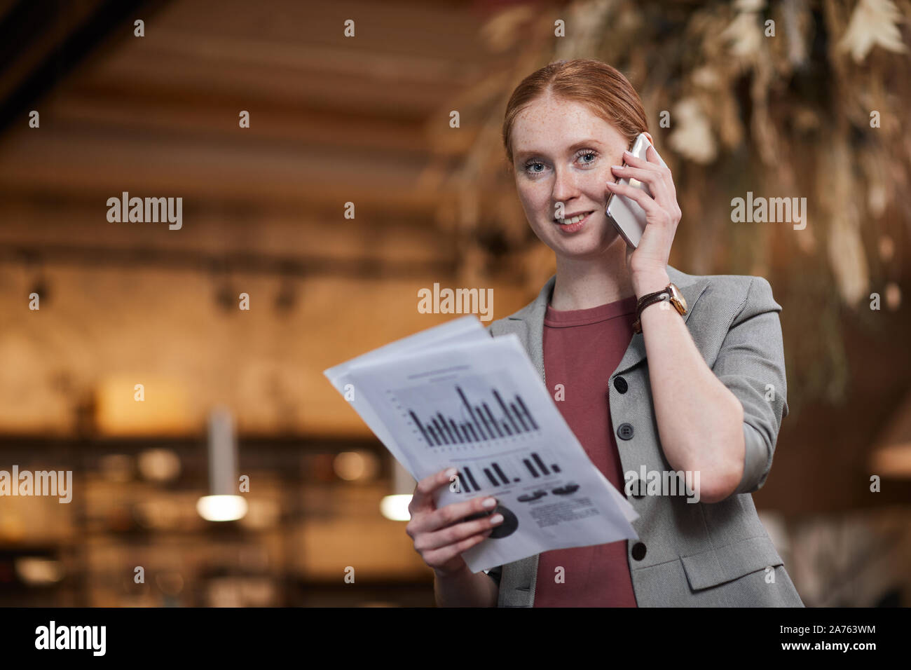 Portrait of red haired businesswoman standing and smiling at camera while has a conversation on mobile phone and examining financial documents Stock Photo