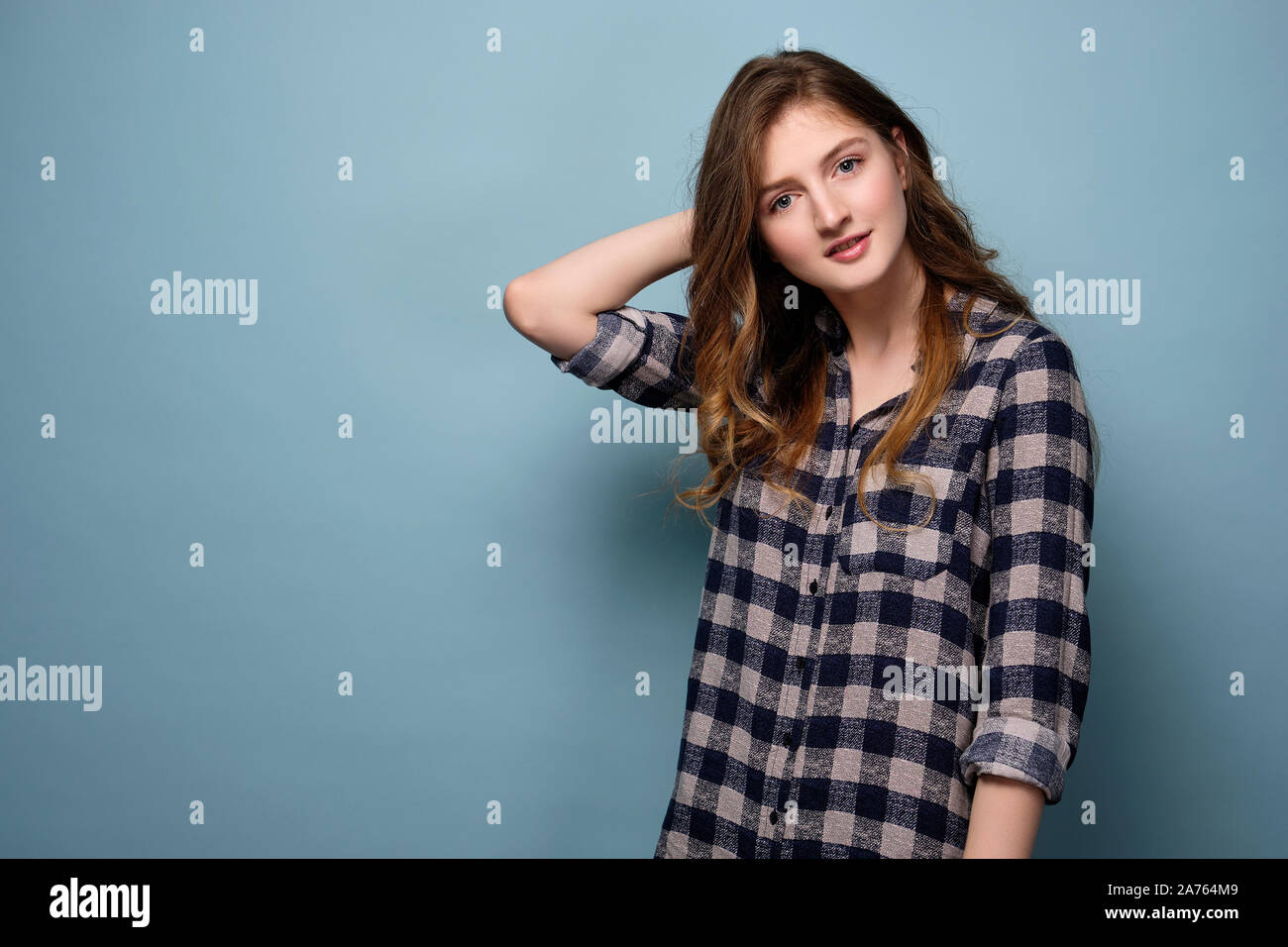 A young girl in a plaid shirt stands on a blue background and looks at the camera with her head bowed, her hand behind her head. Stock Photo
