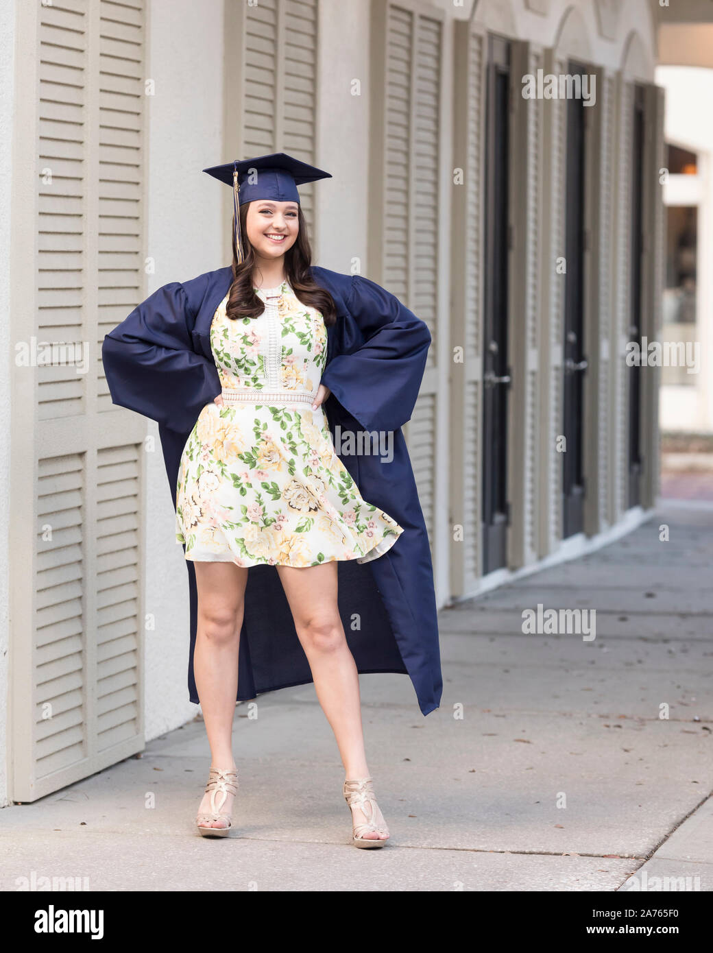 Graduating senior in cap and gown and floral dress poses on street Stock Photo