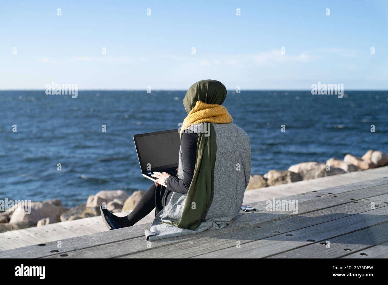 Young woman wearing headscarf sitting beside ocean using laptop Stock Photo