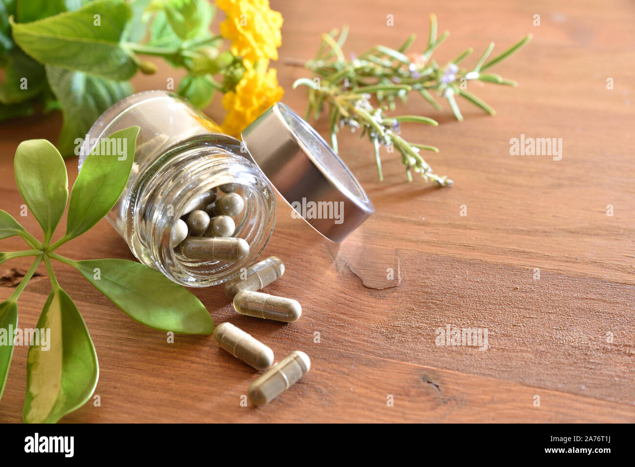 Natural medicine capsules in open glass jar lying on wood table with flowers and plant leaves. Natural medicine concept. Elevated view. Horizontal com Stock Photo
