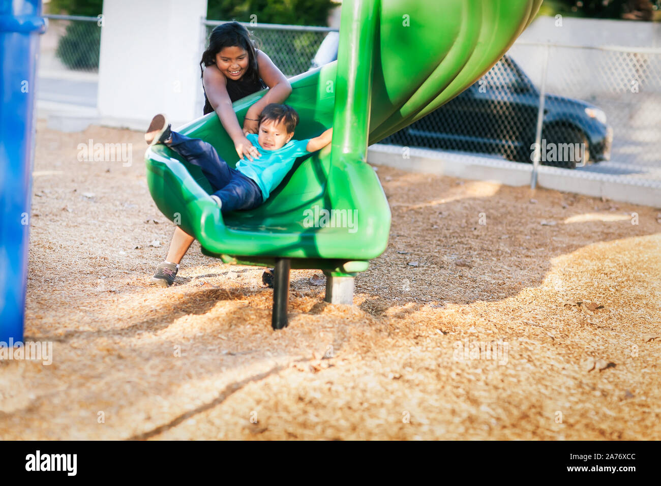 Older sister helping her younger brother go down a twisted slide in a kids playground. Stock Photo