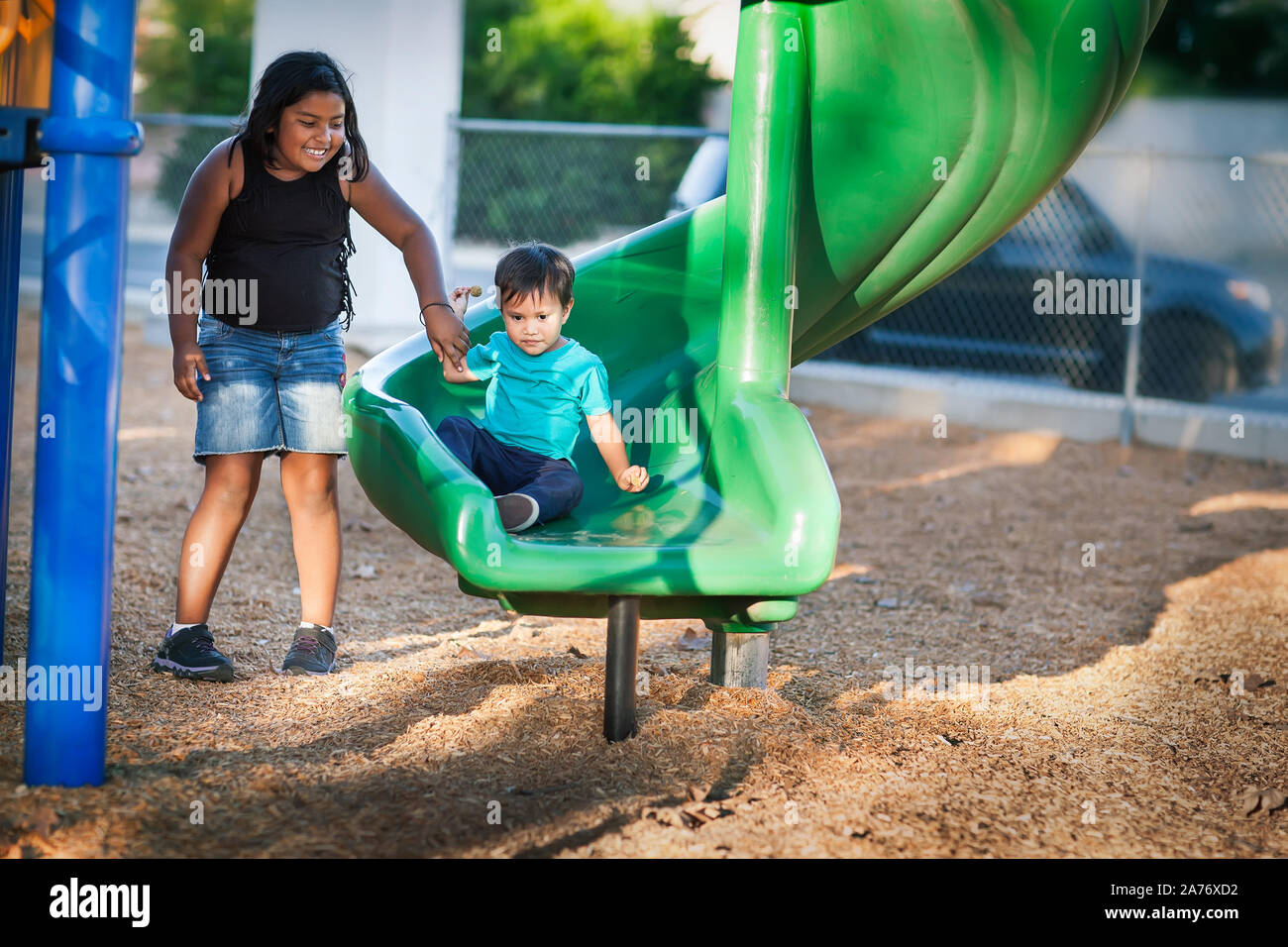 A big sister is holding her young brothers hand while he gets off from a playground slide. Stock Photo