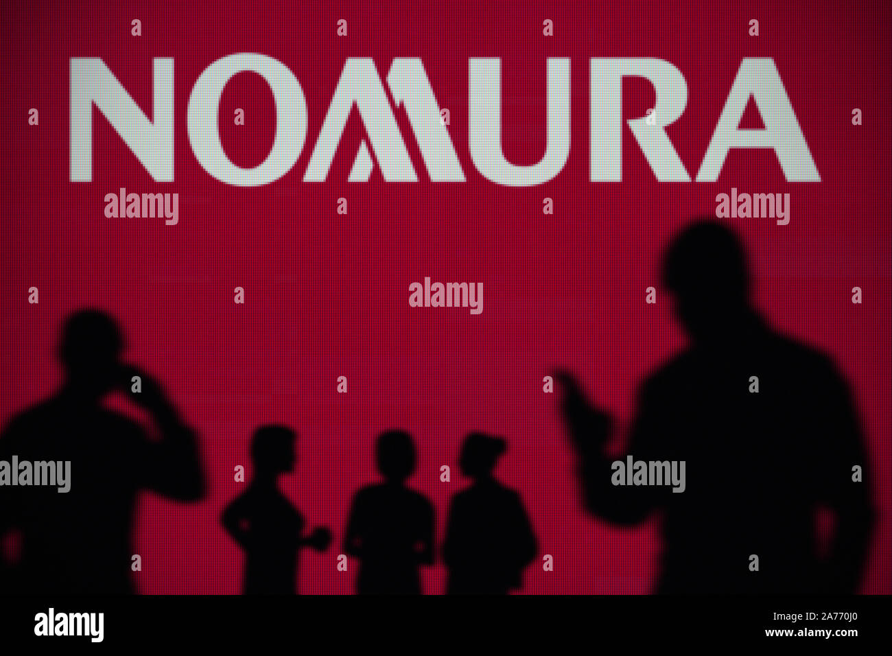 The Nomura logo is seen on an LED screen in the background while a silhouetted person uses a smartphone (Editorial use only) Stock Photo