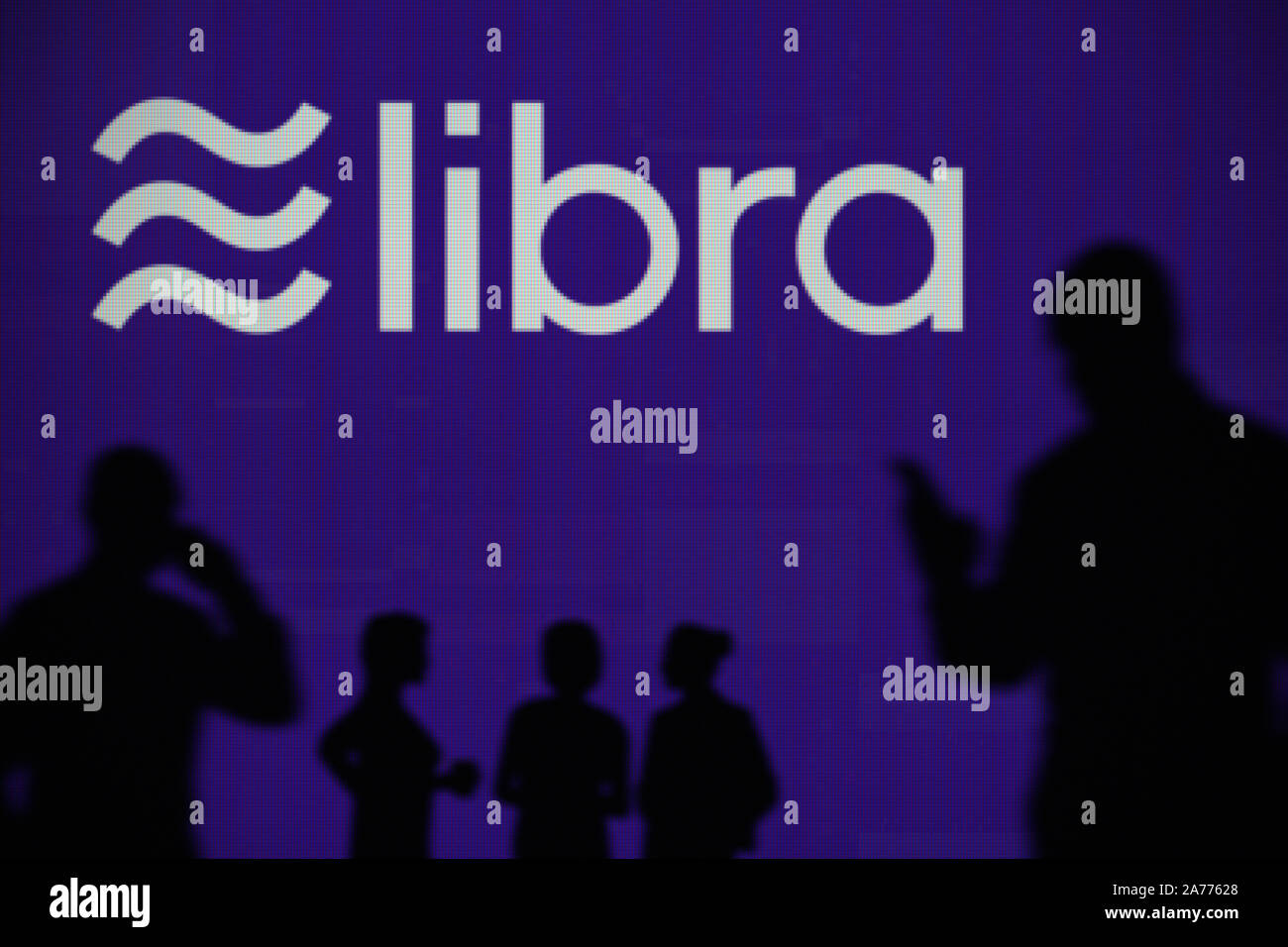 The Libra cryptocurrency logo is seen on an LED screen in the background while a silhouetted person uses a smartphone (Editorial use only) Stock Photo