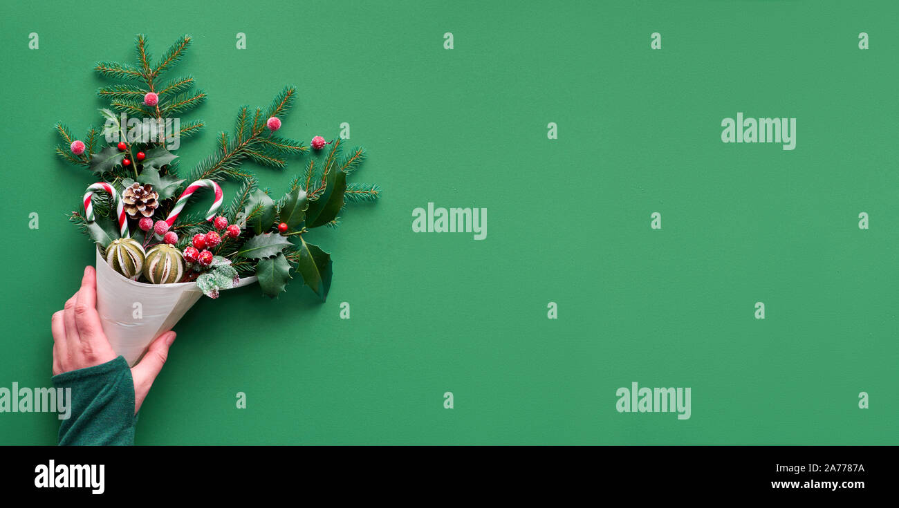 Christmas panoramic background flat lay on green paper with copy-space. Female hand holding veneer cone with fir and holly twigs decorated with candy Stock Photo