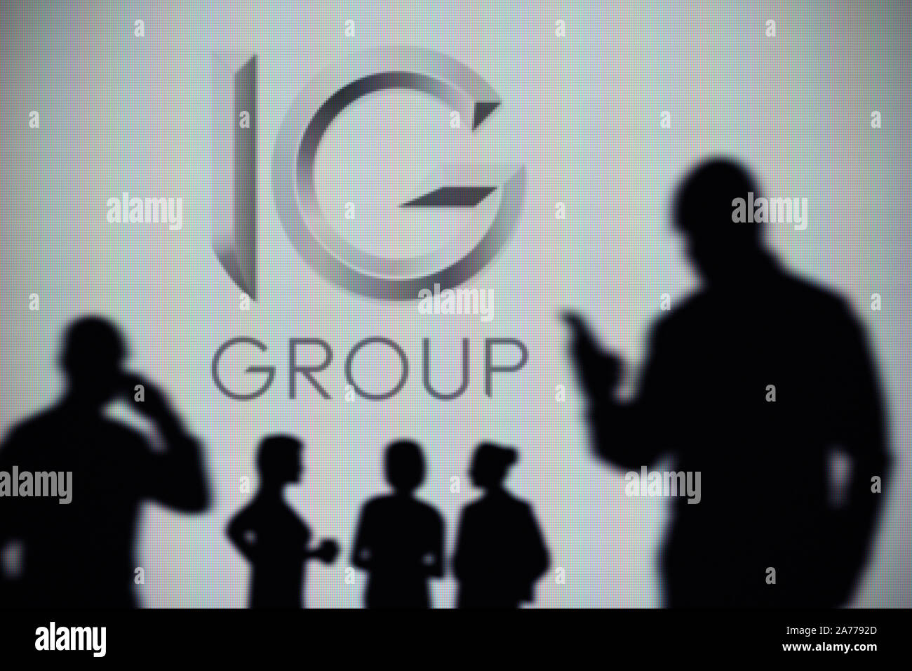 The IG Group logo is seen on an LED screen in the background while a silhouetted person uses a smartphone (Editorial use only) Stock Photo